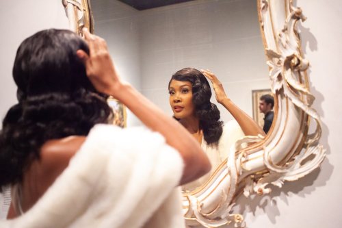 A woman in a white shawl adjusts her hair while looking into an ornate mirror in an art gallery.