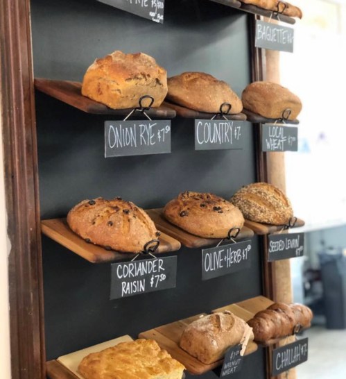 A wooden display shelf with various types of bread, each labeled with prices on small chalkboards.