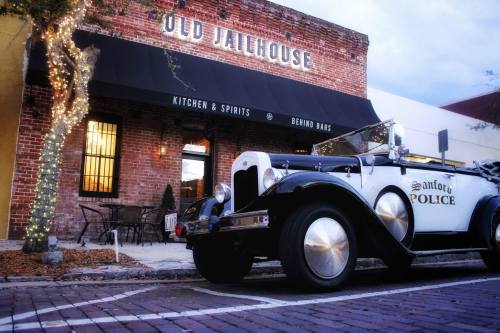 A vintage police car parked in front of the Old Jailhouse restaurant, featuring brick walls and outdoor seating.