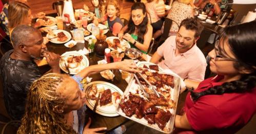 A lively restaurant scene with diners enjoying a meal, as a server offers a platter of ribs to a table.