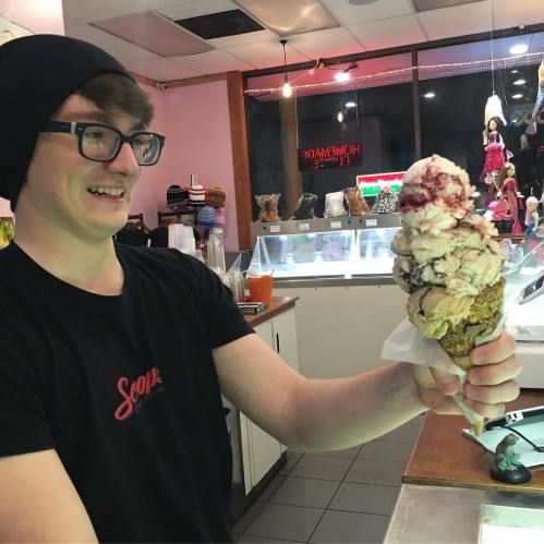 A smiling person in a black beanie holds a towering ice cream cone with multiple scoops in a colorful ice cream shop.
