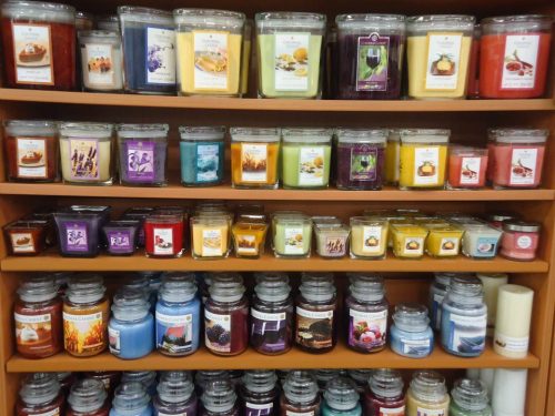 A display of various scented candles in jars, organized by color and scent, on wooden shelves.
