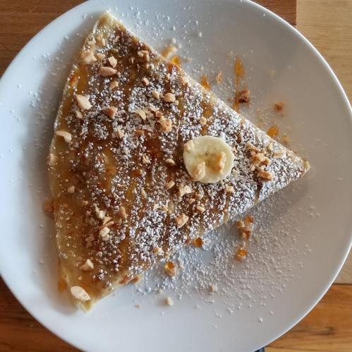 A triangular crepe topped with banana slices, crushed nuts, and powdered sugar on a white plate.