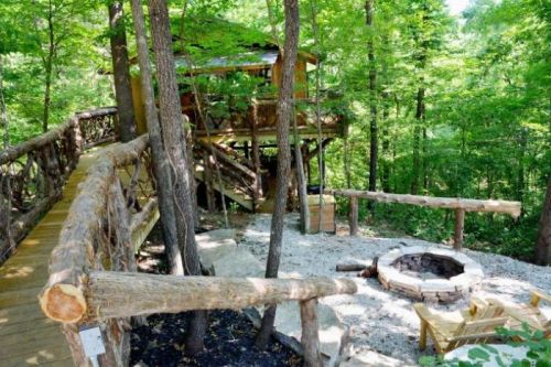 A rustic treehouse surrounded by trees, with a wooden walkway and a stone fire pit in a serene forest setting.