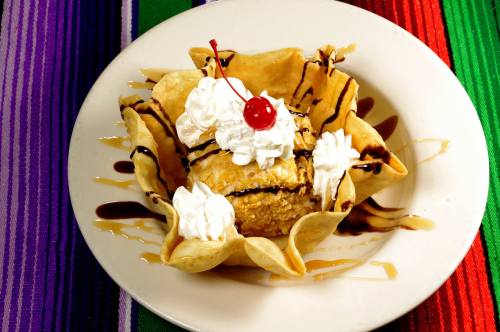 A dessert in a tortilla bowl, featuring ice cream, whipped cream, chocolate drizzle, and a cherry on top.