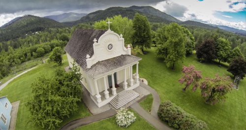 Aerial view of a white church surrounded by lush greenery and mountains under a cloudy sky.