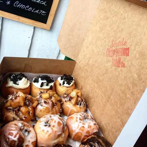 A box of assorted cinnamon rolls, featuring various toppings like cream cheese, chocolate, and apple.
