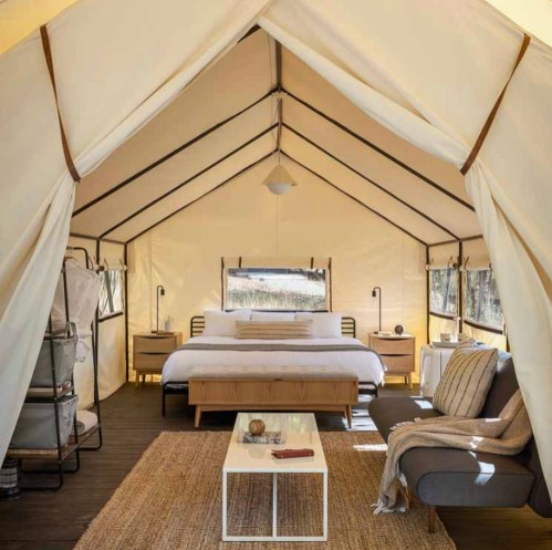 Cozy glamping tent interior with a bed, seating area, and natural light from large windows.