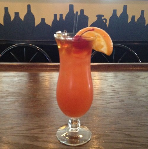 A colorful cocktail in a tall glass, garnished with an orange slice and a cherry, sits on a wooden bar.