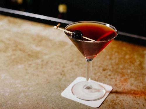 A dark cocktail in a martini glass, garnished with a cherry on a skewer, resting on a coaster.
