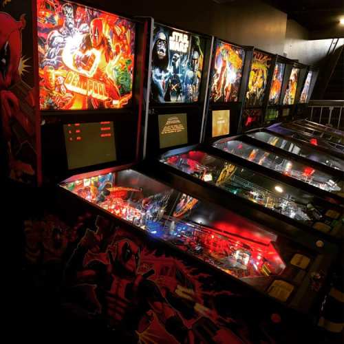 A row of colorful pinball machines featuring various themes, including Deadpool and Star Wars, in a dimly lit arcade.