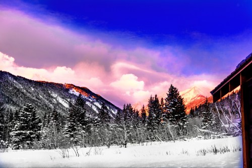 Snow-covered mountains under a vibrant sky at sunset, with evergreen trees in the foreground.