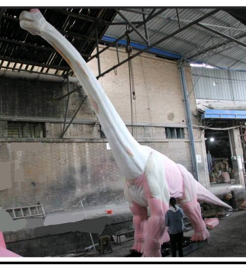 A person stands next to a large, pink and white dinosaur sculpture in a warehouse setting.