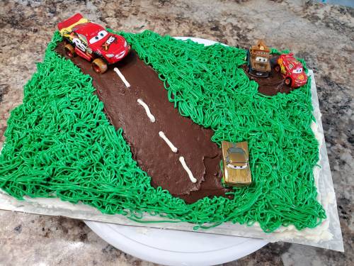 A cake decorated like a racetrack with green frosting, toy cars, and a chocolate road.
