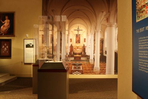 Interior of a museum featuring a display case and a large mural of a church with tall columns and an altar.