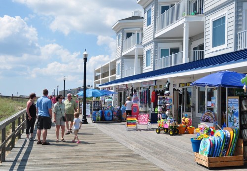 A boardwalk scene with shops selling beach toys, people walking, and colorful umbrellas under a blue sky.