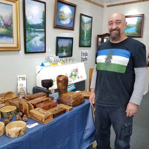 A man stands beside a table displaying wooden crafts and paintings in an art gallery.