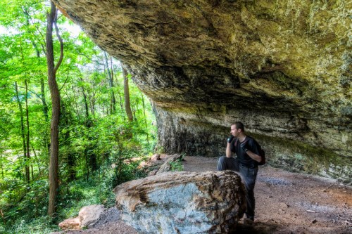 A hiker pauses thoughtfully near a large rock under a rocky overhang, surrounded by lush green trees.