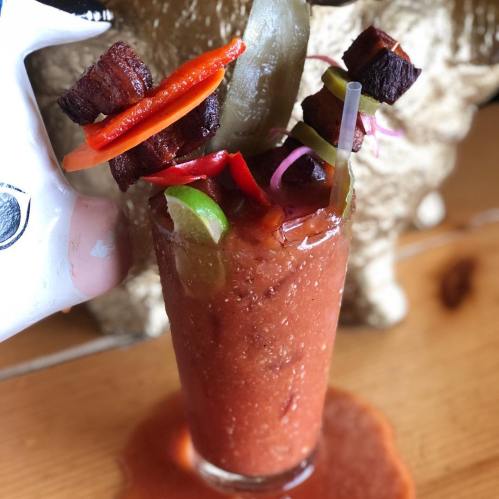 A vibrant Bloody Mary garnished with skewers of meat, peppers, and pickles, with a splash on the table.