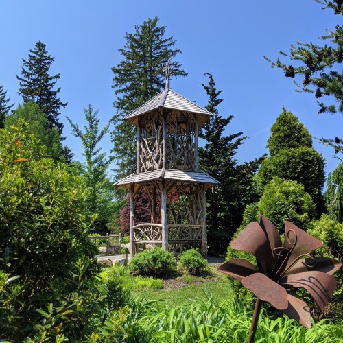 A wooden gazebo surrounded by lush greenery and trees, with a metal flower sculpture in the foreground.