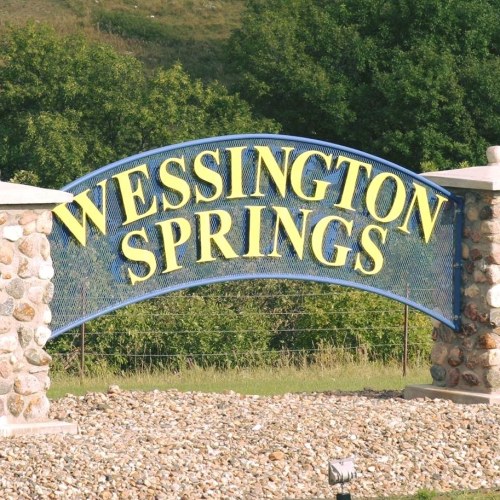 Sign for Wessington Springs, featuring bold yellow letters on a blue arch, surrounded by stone pillars and greenery.