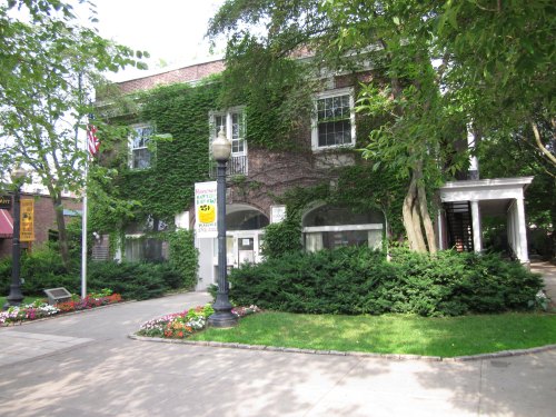 A brick building covered in green ivy, surrounded by trees and colorful flower beds, with a lamppost in front.
