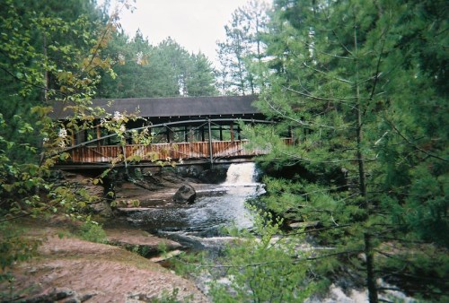 A wooden covered bridge spans a river, surrounded by lush green trees and a small waterfall.