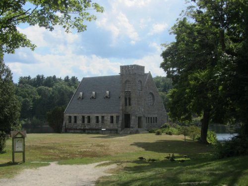 A stone building with a tower, surrounded by trees and a grassy area near a lake under a partly cloudy sky.