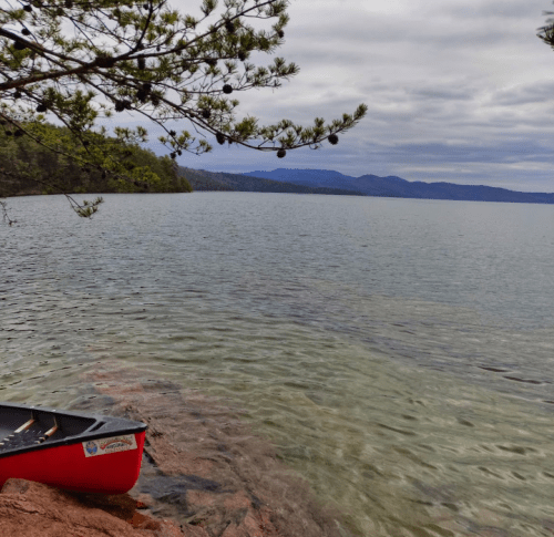 A red canoe rests on a rocky shore by a calm lake, surrounded by trees and distant mountains under a cloudy sky.