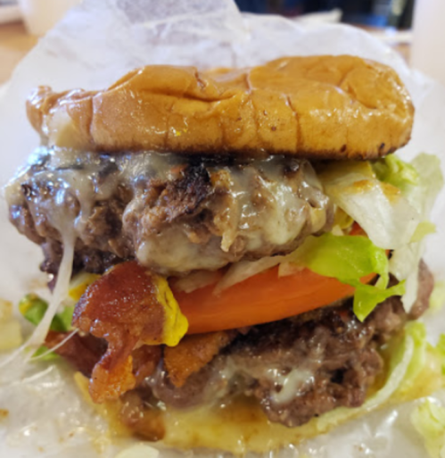 A close-up of a juicy double cheeseburger with bacon, lettuce, tomato, and melted cheese, served in a paper wrapper.