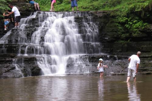 A waterfall cascades into a pool where people, including a child, play and explore the water's edge.