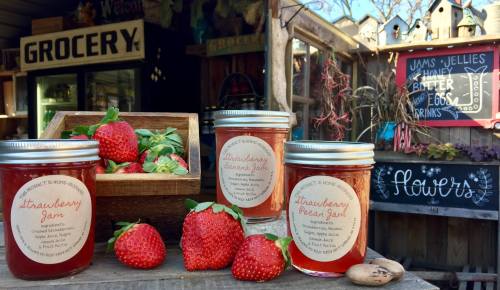 Three jars of strawberry jam and pear jam are displayed with fresh strawberries in a rustic grocery setting.