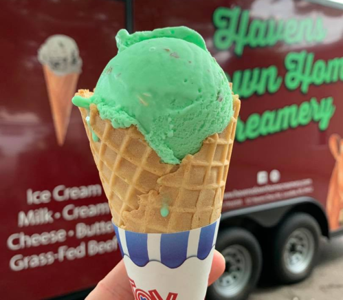 A hand holding a cone with bright green mint ice cream in front of a creamery truck.