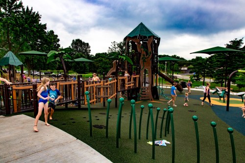 Children play on a colorful playground with a large climbing structure and green landscaping under a cloudy sky.