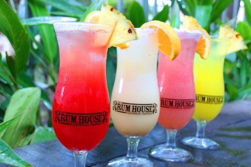 Four colorful cocktails in tall glasses, garnished with pineapple and orange slices, set against a lush green background.