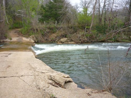 A serene river flows over a rocky ledge, surrounded by lush greenery and trees on a cloudy day.