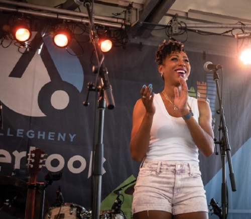 A smiling performer in a white tank top and shorts claps on stage, with microphones and instruments in the background.