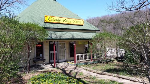 A quaint bakery with a green roof and "Serenity Farm Bread" sign, surrounded by trees and blooming flowers.