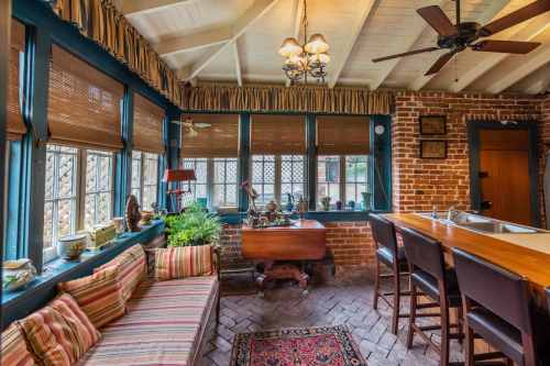 Cozy kitchen with brick walls, large windows, a wooden table, and a striped sofa, featuring plants and decorative items.