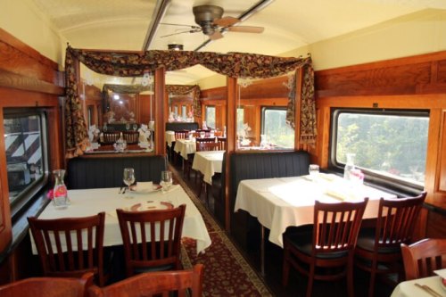 Interior of a vintage dining car with elegant tables, chairs, and large windows, featuring floral curtains and a cozy atmosphere.