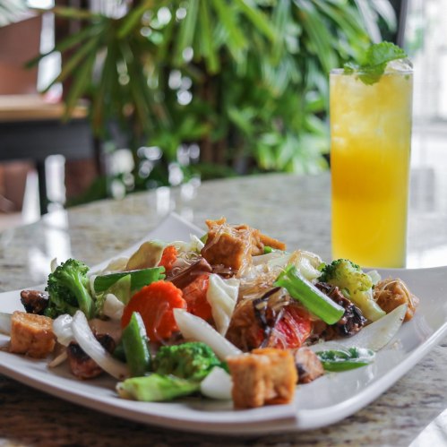 A colorful plate of stir-fried vegetables and tofu, accompanied by a refreshing yellow drink, set against a lush background.