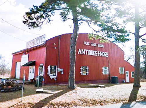 Red barn-style building with "Old Sale Barn Antiques + More" sign, surrounded by trees and a gravel path.