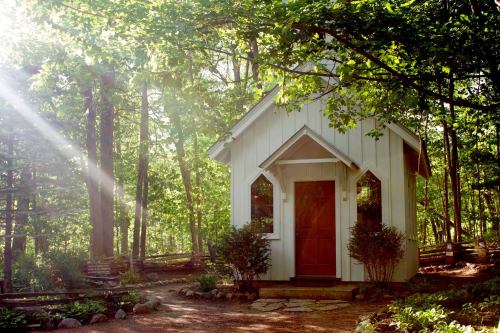 A small white chapel nestled in a lush green forest, surrounded by trees and sunlight filtering through the leaves.