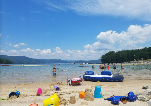 A sunny beach scene with people swimming, colorful sand toys scattered, and a blue inflatable raft on the water.
