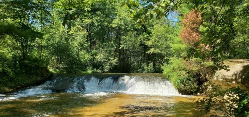 A serene waterfall cascades over rocks, surrounded by lush green trees and a clear blue sky.