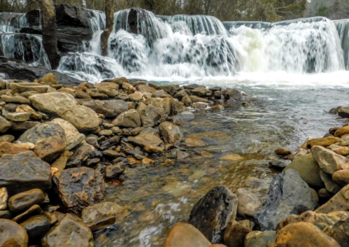A serene waterfall cascades over rocks, surrounded by a tranquil river and smooth stones along the water's edge.