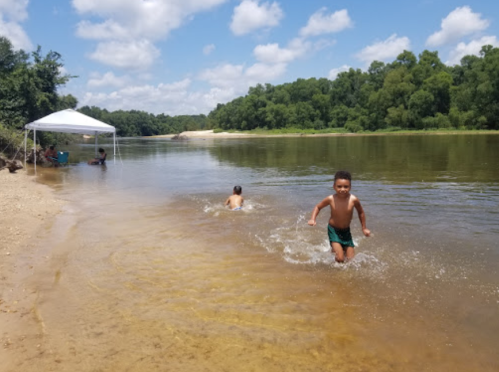 Two children play in a river on a sunny day, with a tent set up on the shore and lush greenery in the background.