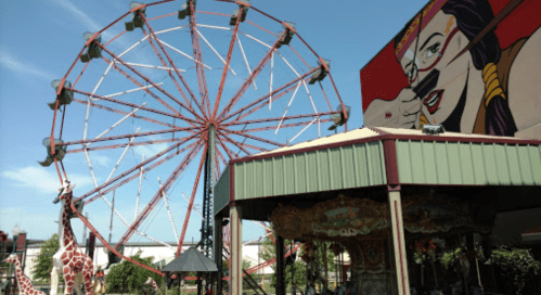 A colorful ferris wheel beside a carousel and giraffe sculptures, with a mural in the background.