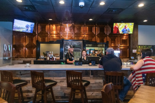 A bar scene with two staff members at the counter and patrons seated, surrounded by wooden decor and TVs.