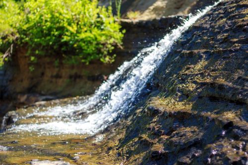 A small waterfall cascades over rocky terrain, surrounded by lush green foliage under bright sunlight.